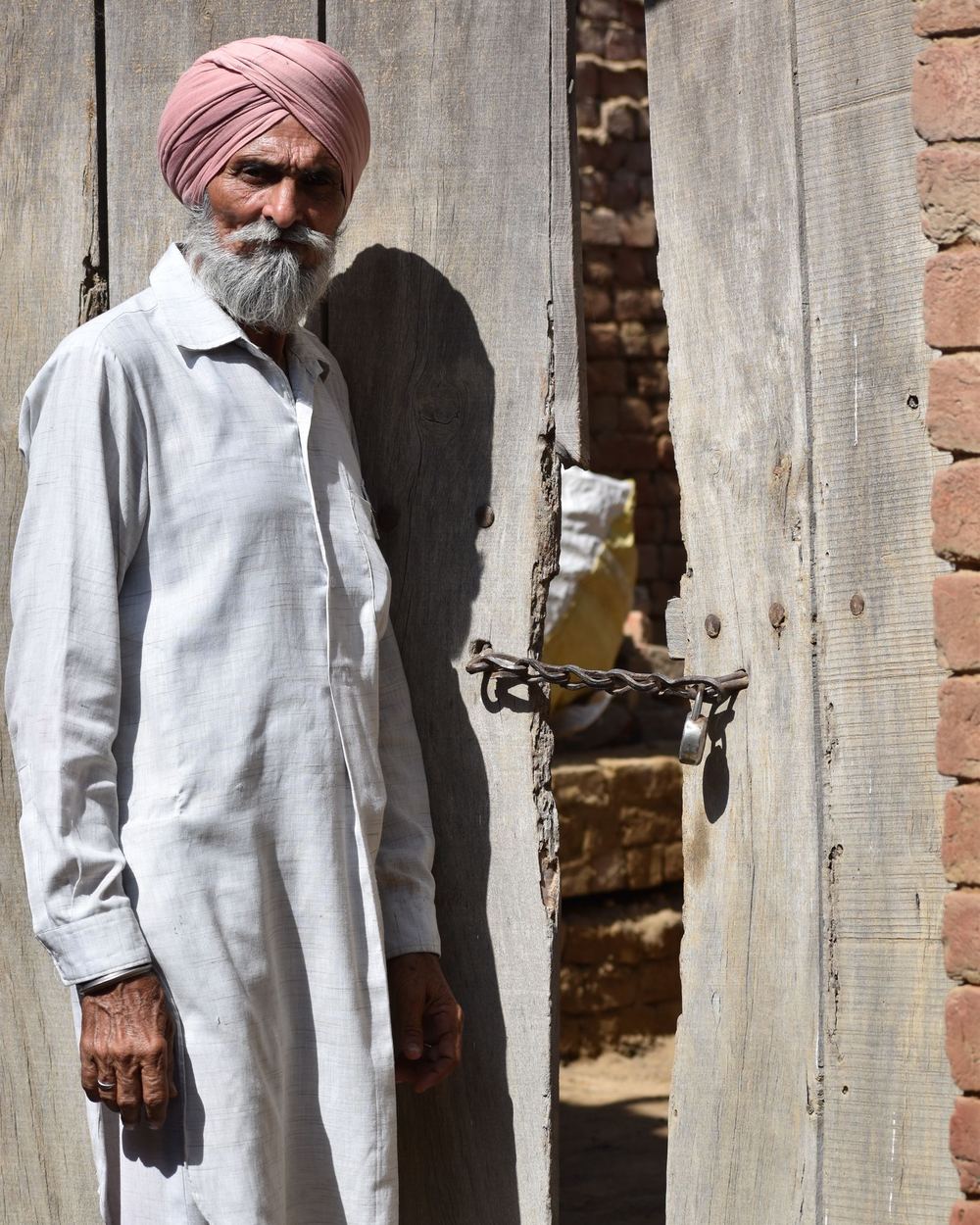 An elder in a turban standing by a wooden gate in a Punjab village
